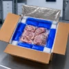 Open insulated shipping box filled with frozen meat, blue gel packs and white cooling packs in a cold-storage facility.